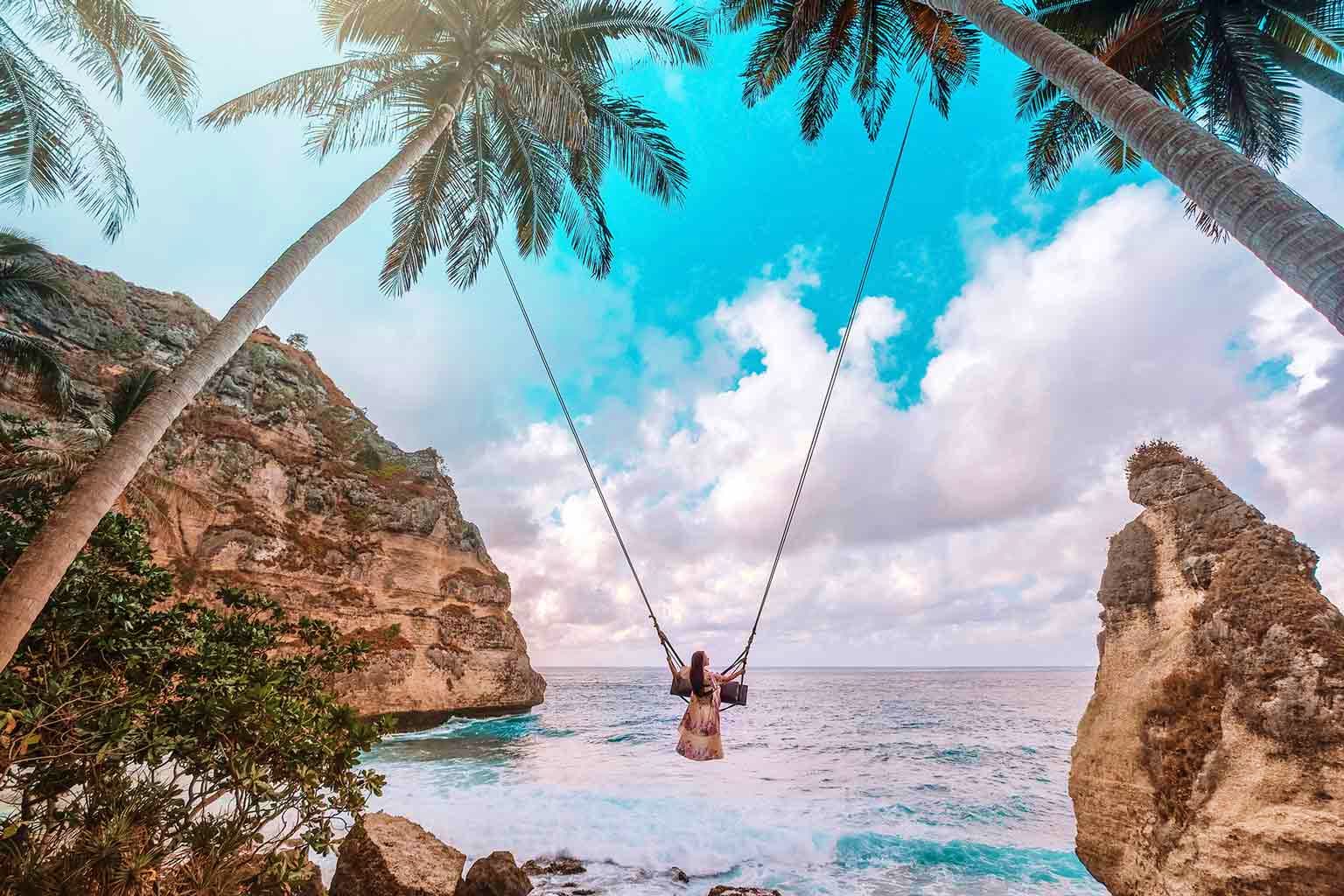 Indonesia Visa Woman on swing hanging from palm tree over turquoise ocean between two limestone rock formations in Bali, Indonesia